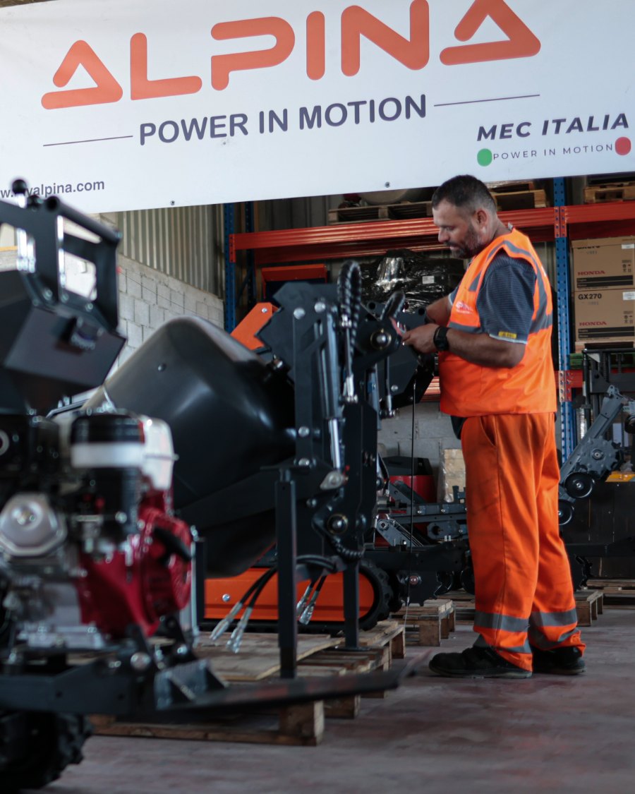 Il capannone MEC Italia: macchine Alpina in fila sui pallet sotto l'insegna, un operaio al lavoro in fondo
